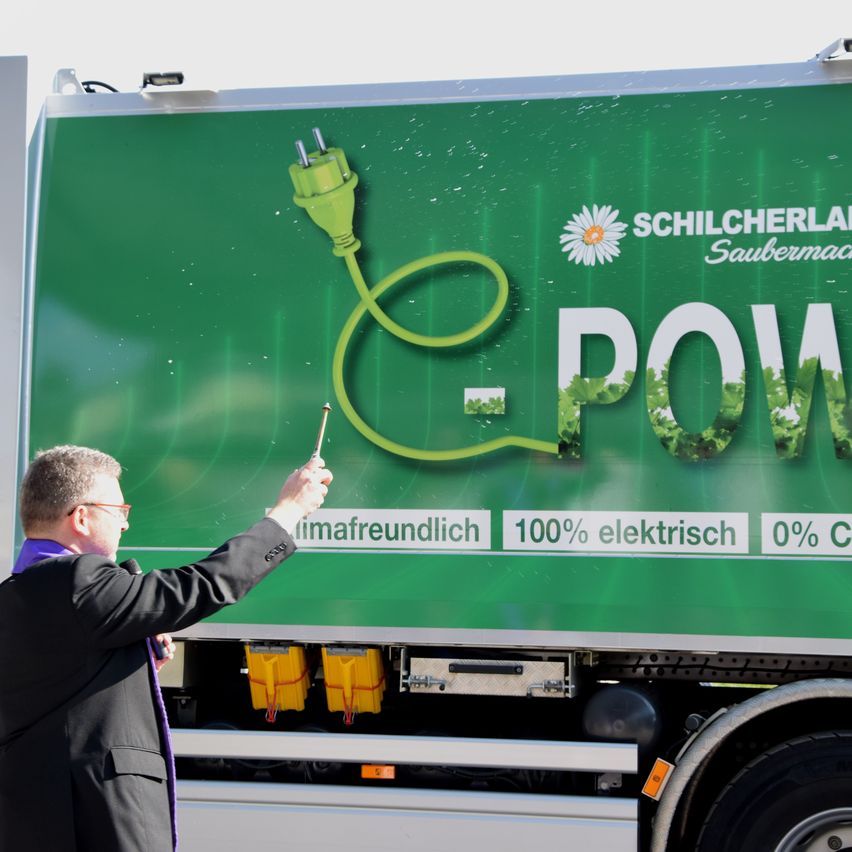 Ein Mann zeigt auf eine grüne Werbung auf einem LKW. Sie zeigt einen Stecker, grüne Blattdarstellungen und den Slogan -POWER. Die Anzeige lautet klimafreundlich, 100% elektrisch, 0% C.