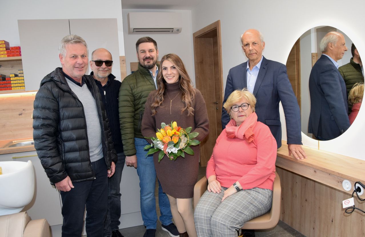 Fünf Erwachsene stehen und sitzen in einem Raum. Eine Frau in einem braunen Kleid hält einen Blumenstrauß. Eine andere Frau sitzt in einem Stuhl und trägt eine Brille. Hinter ihnen steht ein Mann, der lächelt, und ein anderer Mann steht mit einer Sonnenbrille. Ein Spiegel reflektiert sie.
