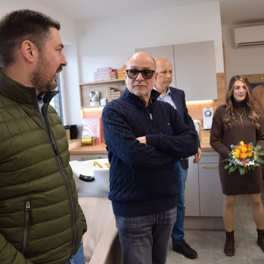 Eine Gruppe von vier Personen in einer Küche, ein Mann in einer grünen Jacke, ein weiterer mit einer Brille, eine Frau mit einem Blumenstrauß und ein Mann im Anzug.