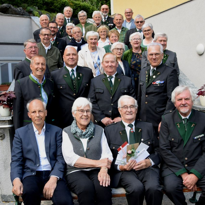 Eine Gruppe von Menschen in Uniformen posiert für ein Foto auf einer Steintreppe, einige halten Bücher und Blumen. Im Hintergrund befinden sich Pflanzen und eine Wand.