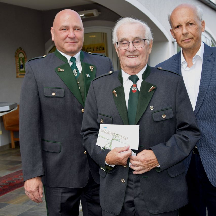 Drei Männer in formeller Kleidung stehen zusammen, einer hält ein Zertifikat, vor einem Gebäude.