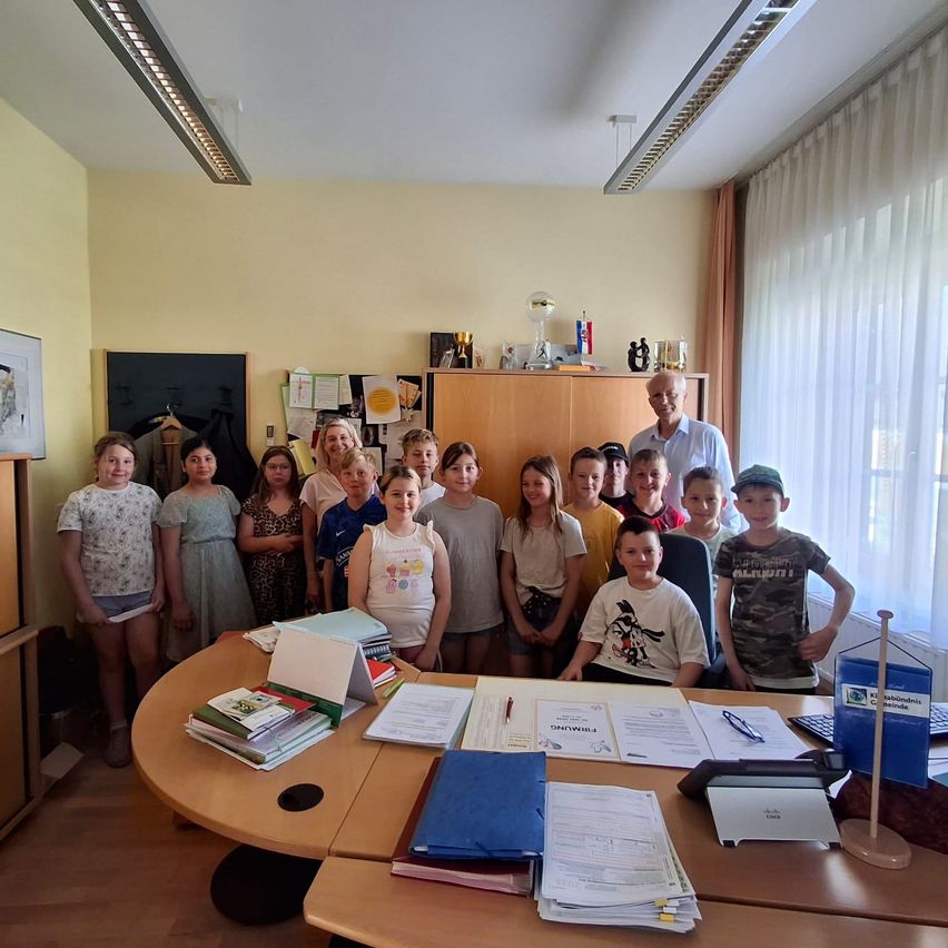 Eine Gruppe von Kindern und Erwachsenen steht um einen großen Schreibtisch mit Papieren, Büchern und Büromaterial in einem Raum mit gelben Wänden und weißen Vorhängen.