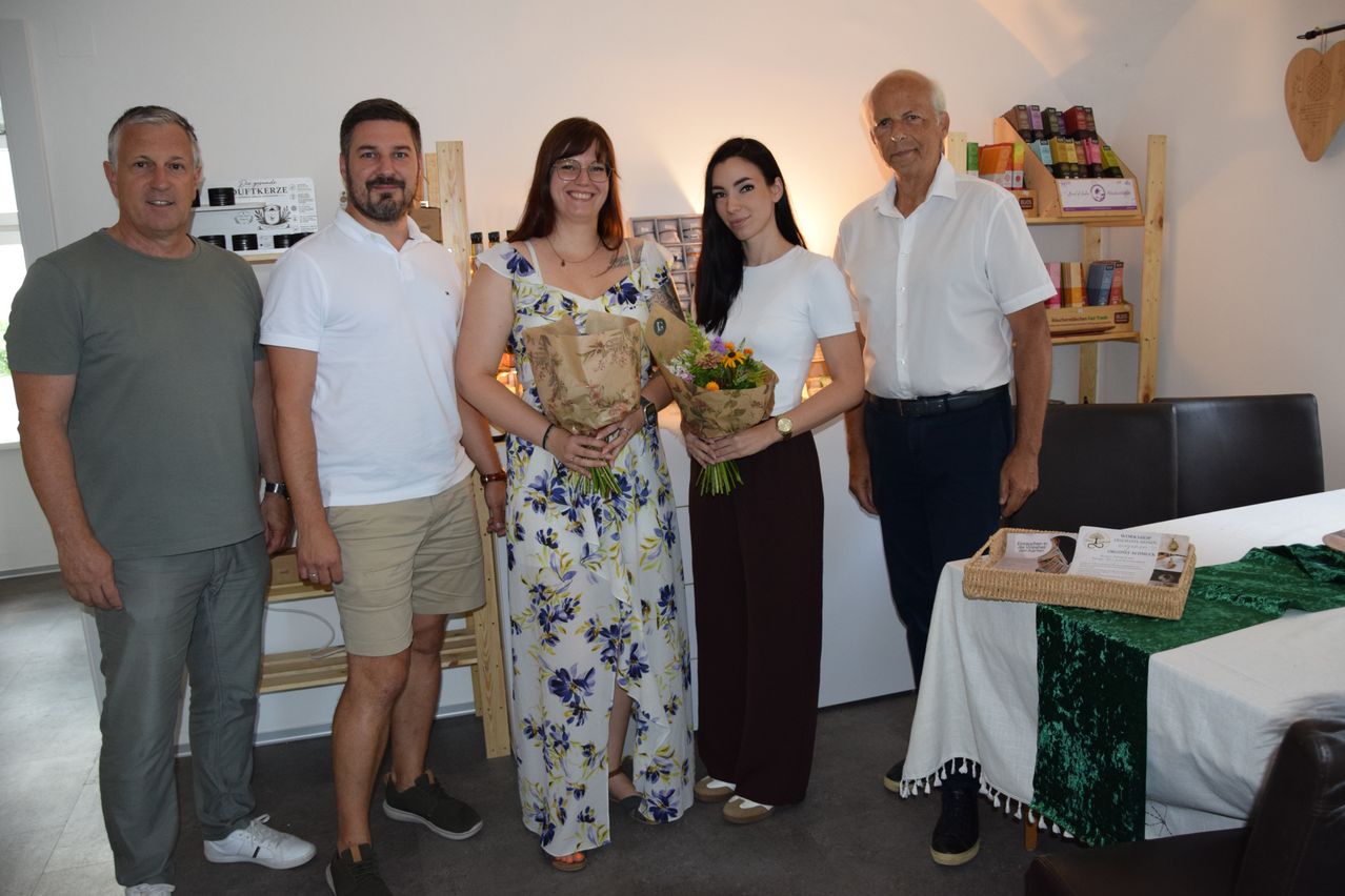 Fünf Personen stehen in einem Raum, zwei Frauen halten Blumensträuße, ein Mann im Anzug und zwei Männer in Freizeitkleidung. Sie scheinen für ein Foto zu posieren.