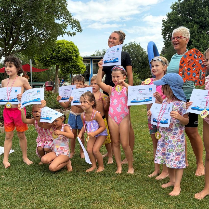 Eine Gruppe von Kindern und Erwachsenen steht auf einem Rasen, hält Urkunden und Medaillen in den Händen. Sie lächeln und posieren für ein Foto. Die Kinder tragen Badekleidung und einige von ihnen haben Schwimmbrillen. Die Erwachsenen stehen hinter ihnen, und einer von ihnen hält eine Urkunde. Im Hintergrund befinden sich Bäume und ein Gebäude.
