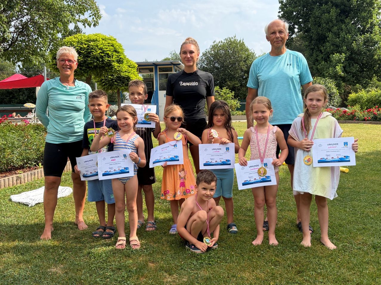Eine Gruppe von Kindern und Erwachsenen steht auf einem Rasenfeld, hält Urkunden und Medaillen in den Händen. Sie alle lächeln und posieren für ein Foto. Die Kinder tragen Badekleidung und die Erwachsenen tragen Freizeitkleidung. Hinter ihnen stehen Bäume und ein Gebäude.