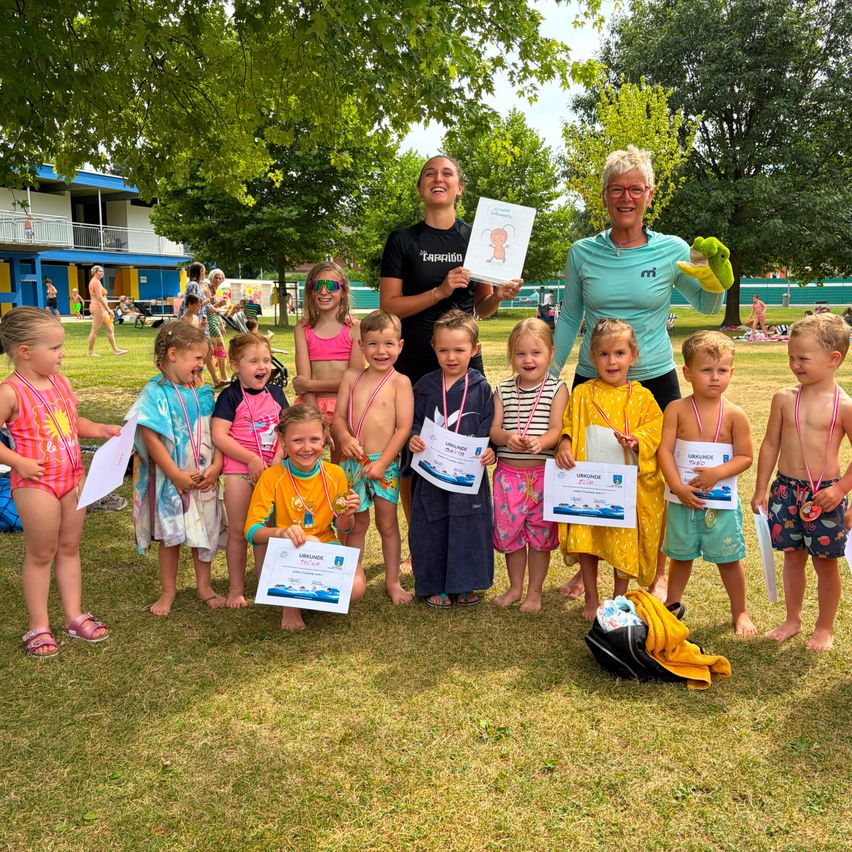 Eine Gruppe von Kindern und zwei Erwachsenen steht auf einem Rasenfeld, hält Urkunden und lächelt. Sie sind von Bäumen und einem Gebäude im Hintergrund umgeben.