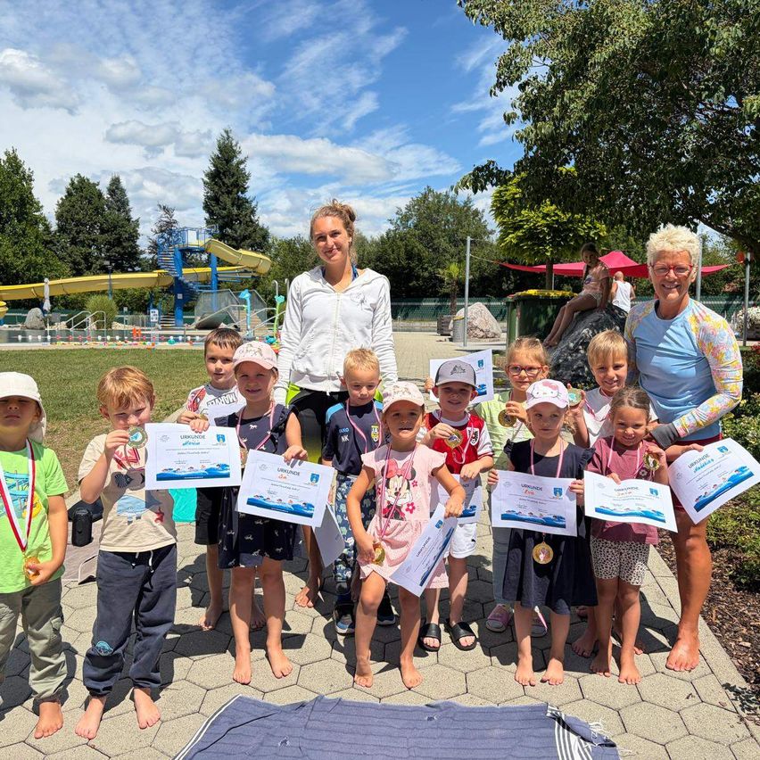 Eine Gruppe von Kindern und zwei Erwachsenen steht in einem Park, hält Urkunden und Medaillen in der Hand, mit einem Wasserpark im Hintergrund.