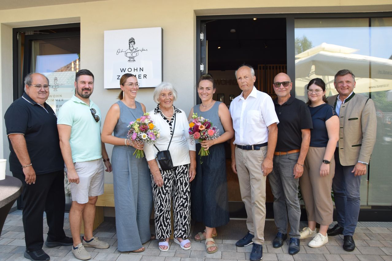 Eine Gruppe von Menschen, darunter zwei Frauen mit Blumensträußen, steht vor einem Gebäude mit einem Schild, das 'Wohn Zimmer' besagt.