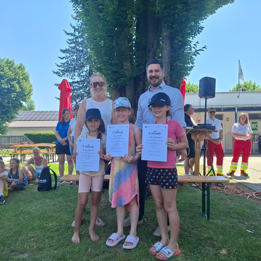 Eine Familie mit drei Kindern steht unter einem Baum und hält Urkunden in der Hand. Sie sind von anderen Menschen umgeben, möglicherweise bei einer Veranstaltung im Freien. Im Hintergrund befinden sich Bänke und ein Gebäude mit Solarmodulen.