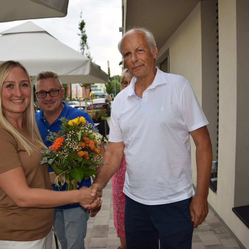 Eine Frau übergibt einem älteren Mann einen Blumenstrauß im Freien. Zwei weitere Personen stehen hinter ihnen in der Nähe eines weißen Regenschirms.