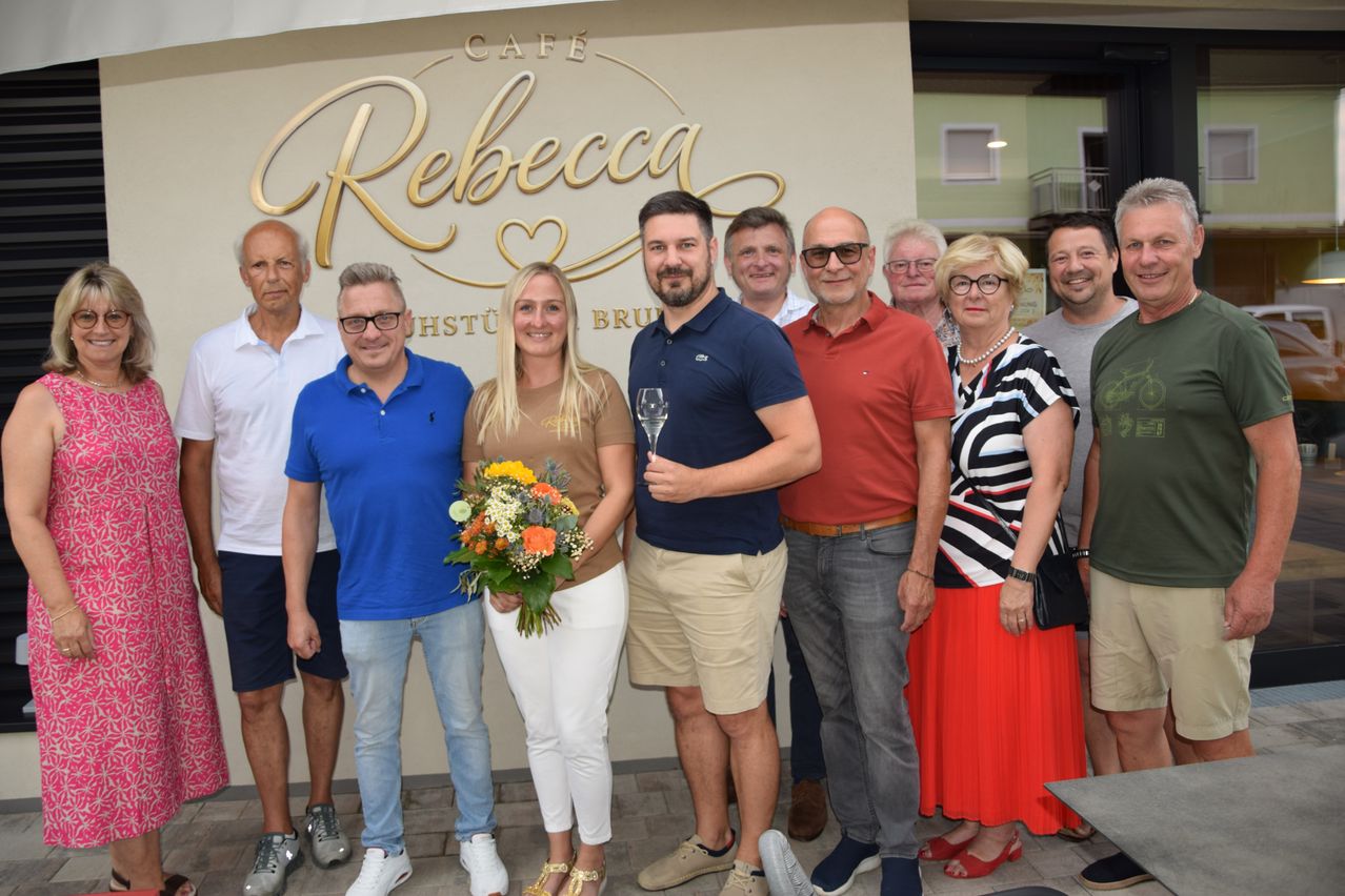 Eine Gruppe von Menschen steht vor dem Cafe Rebecca, lächelt und hält ein Weinglas und einen Blumenstrauß. Sie sind lässig gekleidet und scheinen ein Ereignis zu feiern.