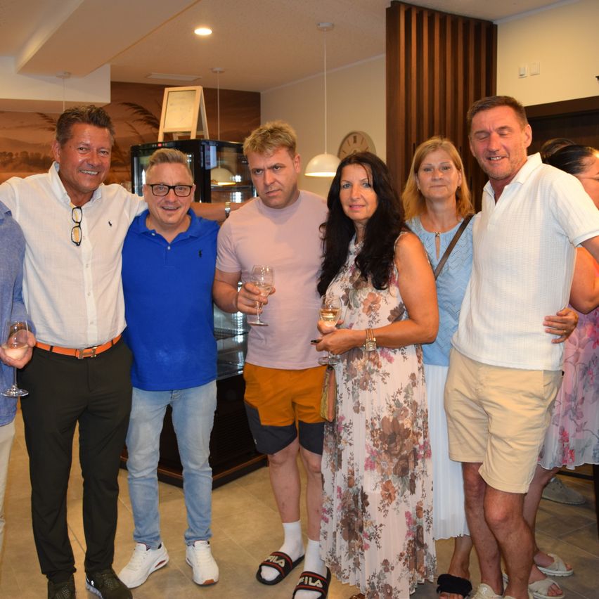 Eine Gruppe von Menschen in heller Kleidung posiert für ein Foto mit Weinglas in der Hand. Der Mann in der Mitte trägt eine Brille und ein blaues Shirt, während andere Pink, Weiß und Hellblau tragen.