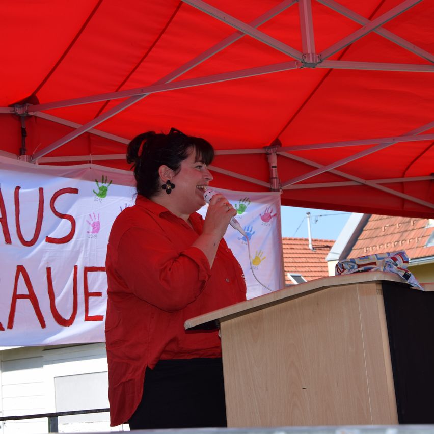 Eine Frau mit kurzen Haaren, die ein rotes Langarmshirt trägt, spricht in ein Mikrofon unter einem roten Zelt. Hinter ihr ist ein Banner mit dem Wort 'AUS RAUE' zu sehen.