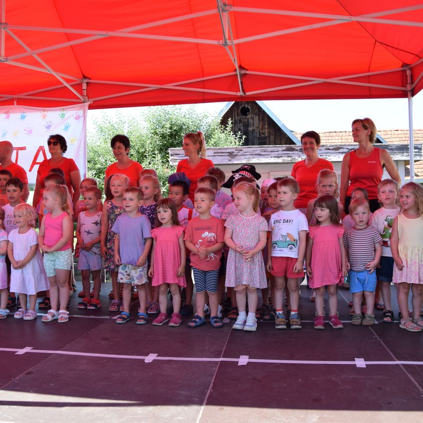 Eine Gruppe von Kindern in roten und rosa T-Shirts steht auf einer Bühne unter einem roten Zelt. Einige tragen Hüte und Turnschuhe. Dahinter stehen vier Frauen in roten Shirts und lächeln.