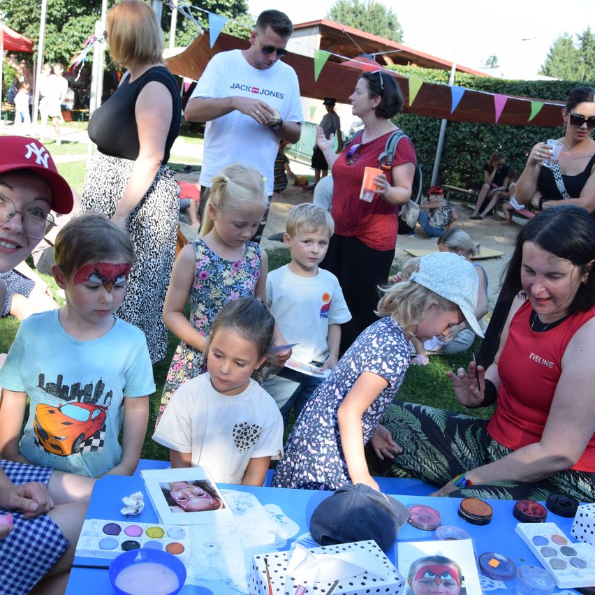 Eine Gruppe von Kindern und Erwachsenen hat eine Gesichtsbemalung-Sitzung bei einer Veranstaltung im Freien. Es gibt Pinseln, Farben und Bilder auf einem Tisch. Erwachsene helfen den Kindern beim Malen.