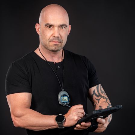Ein Mann mit einem schwarzen T-Shirt, einer Uhr und einem Nackenband hält ein Tablet und schaut darauf. Er hat ein Tattoo auf seinem rechten Arm.