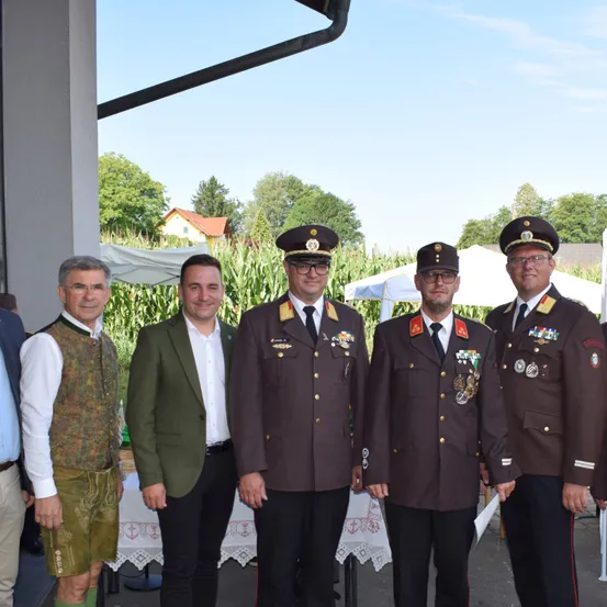 Fünf Männer stehen draußen, gekleidet in verschiedenen Uniformen. Drei Männer tragen Militäruniformen mit Medaillen, während zwei Männer an den Seiten Zivilkleidung tragen. Hinter ihnen steht ein Tisch mit weißer Tischdecke. Bäume und ein Haus sind im Hintergrund zu sehen.