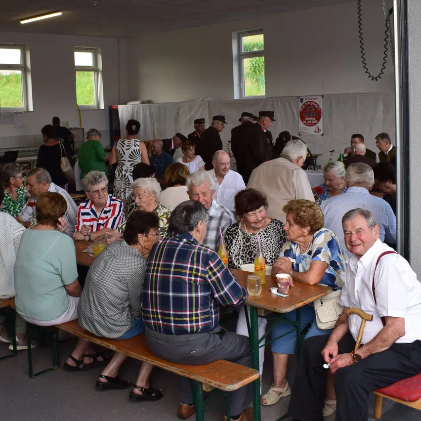 Eine Gruppe älterer Menschen ist in einem Saal versammelt. Einige sitzen an Tischen mit Getränken, während andere stehen und sich unterhalten. Der Saal hat weiße Wände und Glasfenster.