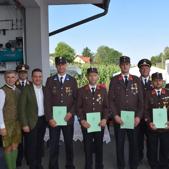 Sieben Männer in Uniform stehen zusammen und halten grüne Urkunden. Dahinter befindet sich ein Maisfeld, ein Gebäude und ein weißes Zelt. Sie feiern wahrscheinlich einen besonderen Anlass.