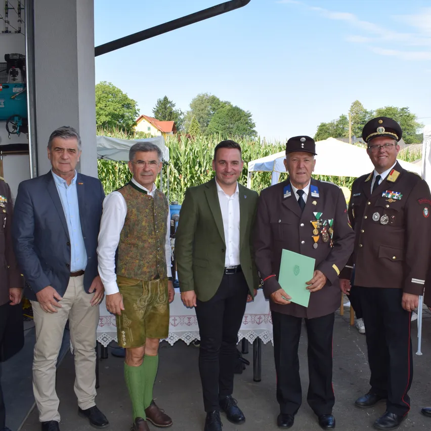 Fünf Männer in Anzügen und Uniformen posieren für ein Foto vor einem Maisfeld. Ein Mann hält eine grüne Mappe. Hinter ihnen steht ein Tisch mit einer weißen Tischdecke.