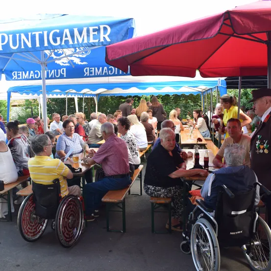 Eine Gruppe älterer Menschen sitzt unter Schirmen in einem Biergarten an Tischen. Einige von ihnen sind im Rollstuhl. Die Schirme sind blau und rot.