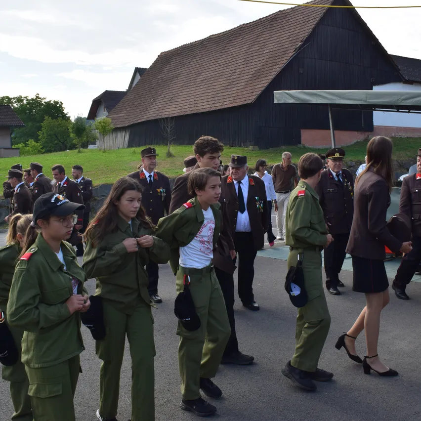 Eine Gruppe uniformierter Personen, möglicherweise Militärkadetten, geht in einer Reihe mit erwachsenen Betreuern auf einem gepflasterten Weg. Im Hintergrund befindet sich ein Haus und ein Baum.