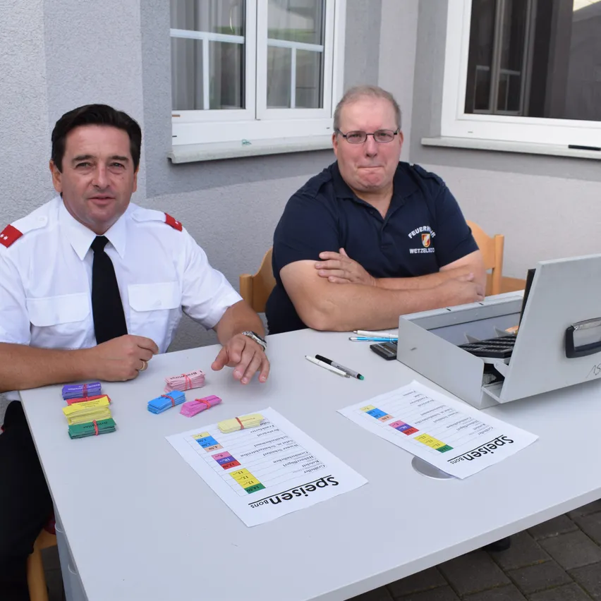 Zwei Männer in Uniformen sitzen an einem Tisch, einer trägt eine Brille, der andere einen Ring. Verschiedene Papiere und eine Box mit dem Wort 'Sicherheit' liegen auf dem Tisch.