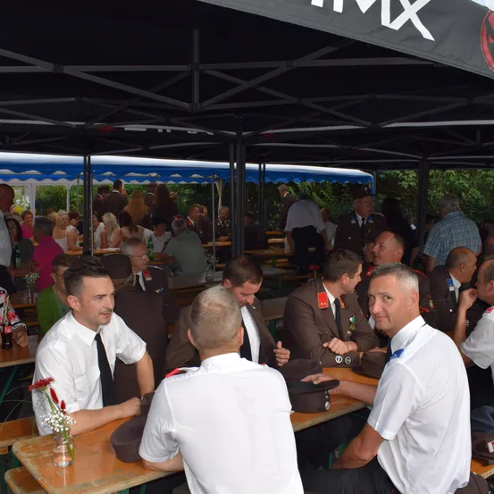 Eine Gruppe von Männern in Uniformen sitzt unter einem Zelt an einem Tisch, möglicherweise bei einer formellen Veranstaltung. Sie sind in ein Gespräch vertieft.