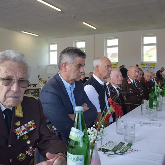 Eine Gruppe von Männern in formeller Kleidung sitzt an einem langen Tisch mit Blumen, Flaschen und Gläsern. Sie scheinen in ein Gespräch vertieft zu sein.