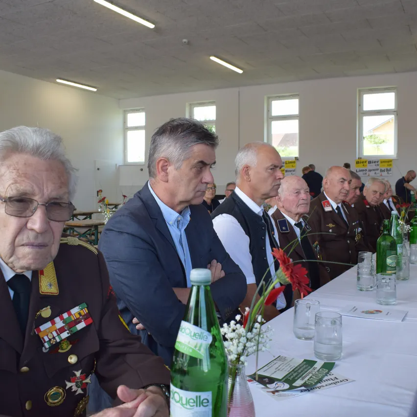 Eine Gruppe von Männern in formeller Kleidung sitzt an einem langen Tisch mit Blumen, Flaschen und Gläsern. Sie scheinen in ein Gespräch vertieft zu sein.