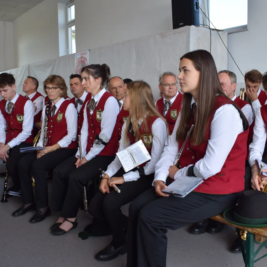 Eine Gruppe von Musikern in roten Westen und schwarzen Hosen sitzt auf einer Bank, einige halten Musikinstrumente, möglicherweise während einer Aufführung.