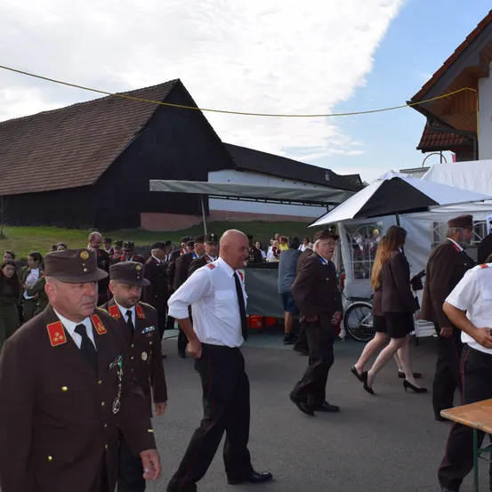 Eine Gruppe von Männern in Uniformen und Mützen geht in einem Außenbereich, möglicherweise bei einer militärischen Veranstaltung. Hinter ihnen stehen Zelte und gehende Menschen, mit einem Scheunen im Hintergrund.