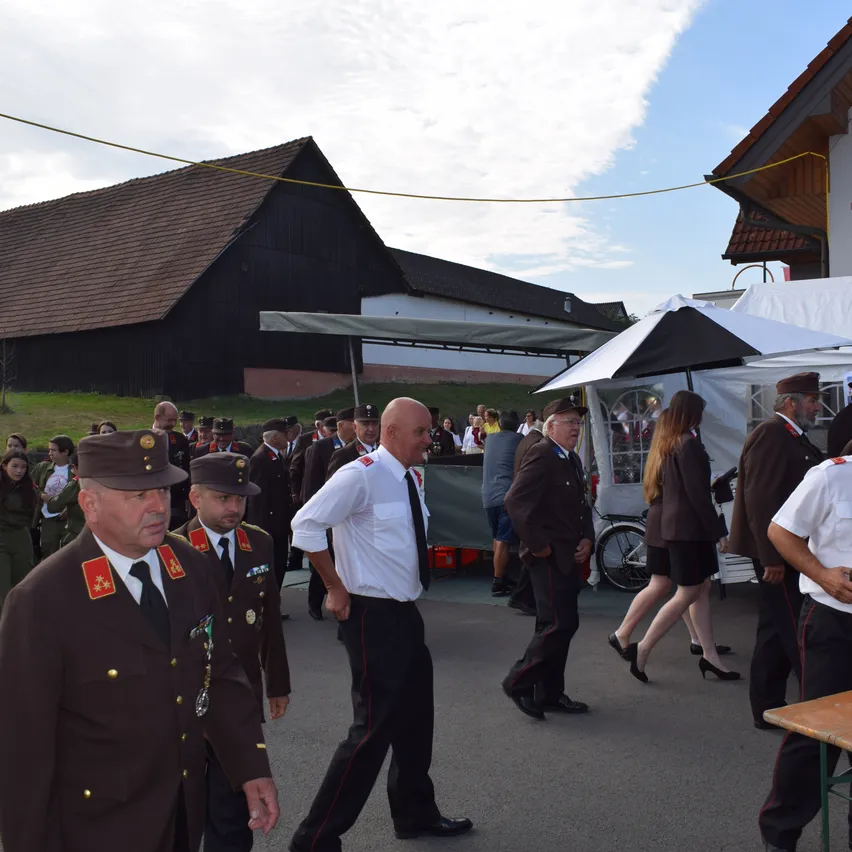 Eine Gruppe von Männern in Uniformen und Mützen geht in einem Außenbereich, möglicherweise bei einer militärischen Veranstaltung. Hinter ihnen stehen Zelte und gehende Menschen, mit einem Scheunen im Hintergrund.