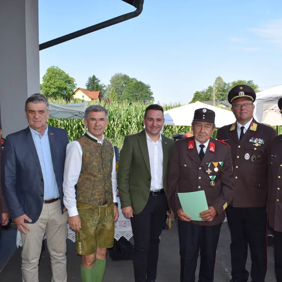 Fünf Männer stehen draußen, einer hält eine Mappe. Zwei tragen Uniformen mit Medaillen, einer in traditioneller Kleidung. Dahinter ein Gebäude und Maisfeld.