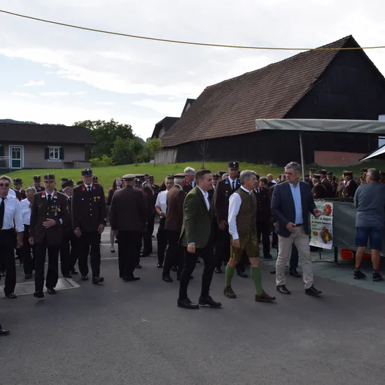 Eine Gruppe formell gekleideter Männer steht vor einem Gebäude. Einige tragen Hüte, Krawatten und Uniformen. Ein anderer Mann hält ein Schild. Sie sind in einem Dorf versammelt.