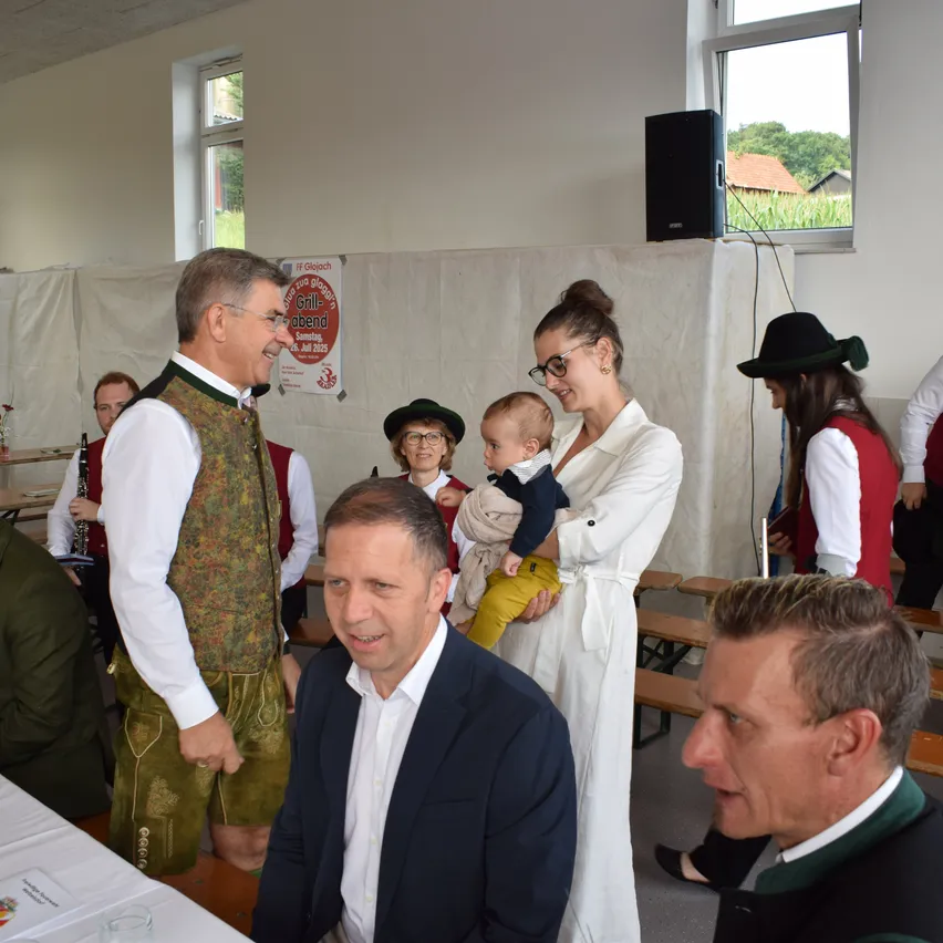 Mehrere Menschen in traditioneller bayerischer Kleidung sind in einem Raum versammelt, darunter ein Mann und eine Frau, die ein Baby halten, ein Mann, der eine Klarinette spielt, und ein Mann in einem schwarzen Anzug, der in die Kamera schaut.