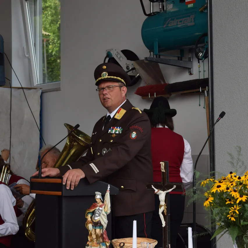 Ein Mann in Uniform hält eine Rede an einem Podium mit Mikrofon und Kreuz dahinter. Eine Statue und Blumen stehen auf dem Podium. Eine weitere Person steht hinter ihm. Eine Tuba befindet sich auf der linken Seite.