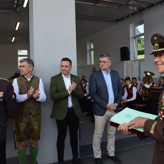 Fünf Männer stehen in einem Innenraum. Ein Mann hält ein Buch, und ein anderer klatscht. Hinter ihnen spielt eine Person die Trompete. Einige Leute sitzen auf dem Boden, und ein Lautsprecher ist rechts aufgestellt.