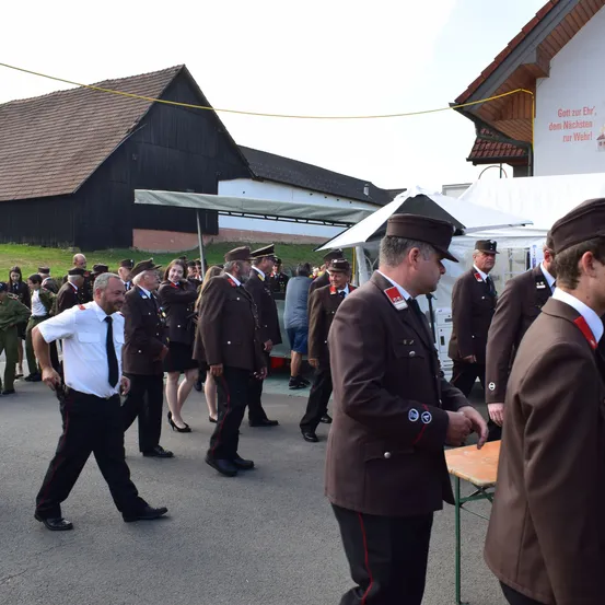 Eine Gruppe von Männern und Frauen in Uniform steht vor einem Gebäude. Einige tragen Hüte, andere sind in Zivilkleidung. Sie scheinen an einer Zeremonie teilzunehmen.