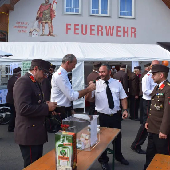 Eine Gruppe von Feuerwehrleuten in Uniform steht um einen Tisch mit einer Schachtel Jägermeister. Dahinter befindet sich ein Gebäude mit einem Wandbild eines Soldaten und dem Wort 'Feuerwehr'.