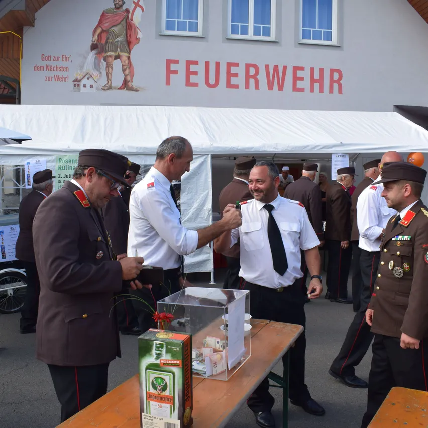 Eine Gruppe von Feuerwehrleuten in Uniform steht um einen Tisch mit einer Schachtel Jägermeister. Dahinter befindet sich ein Gebäude mit einem Wandbild eines Soldaten und dem Wort 'Feuerwehr'.