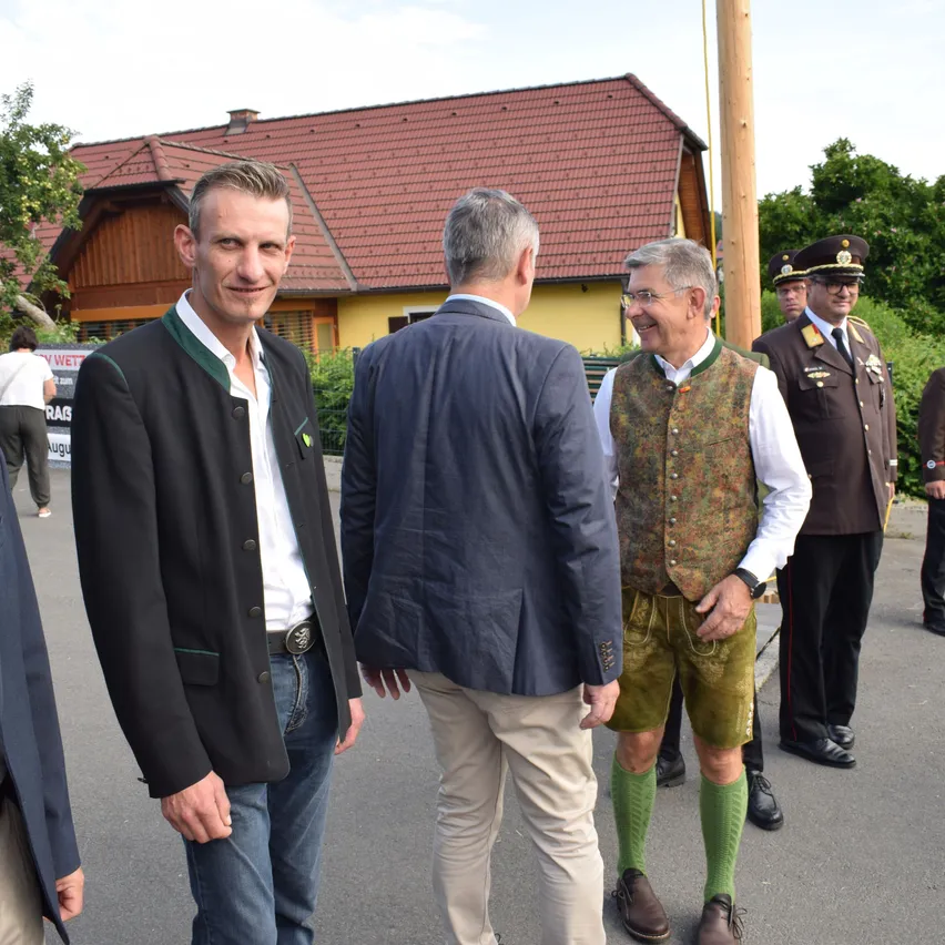 Drei Männer in traditioneller deutscher Kleidung stehen in einem Außenbereich. Dahinter befindet sich ein Haus mit einem roten Dach und einem Pfahl.