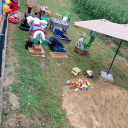 Ein Spielbereich mit Spielzeug auf einem Rasenfeld, darunter eine bunte Elefantenfahrt, ein Piratenschiff und eine Kaktusfahrt. Ein Haufen Spielzeug liegt im Sand. Ein großer Regenschirm steht im Sand.