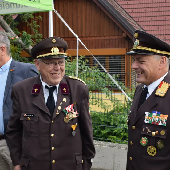 Zwei Männer in Militäruniformen stehen draußen, lächeln und haben einen Zaun und ein Gebäude hinter sich. Ein Mann in einem Anzug beobachtet von links.