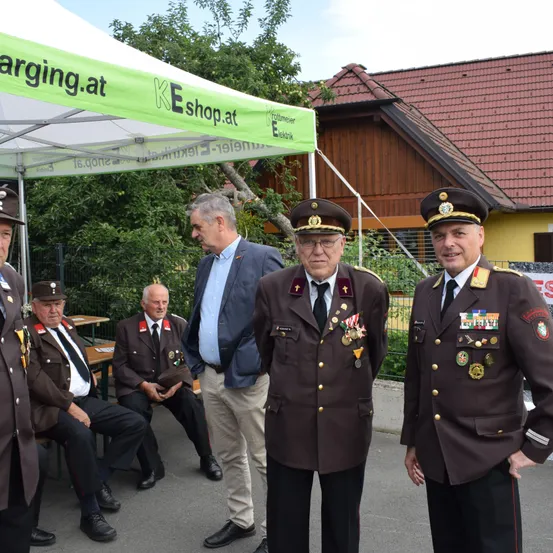 Mehrere Männer in Uniform und ziviler Kleidung stehen vor einem Zelt. Ein Mann in einem Anzug schaut einen sitzenden Mann in Uniform an. Zwei uniformierte Männer stehen neben ihnen.
