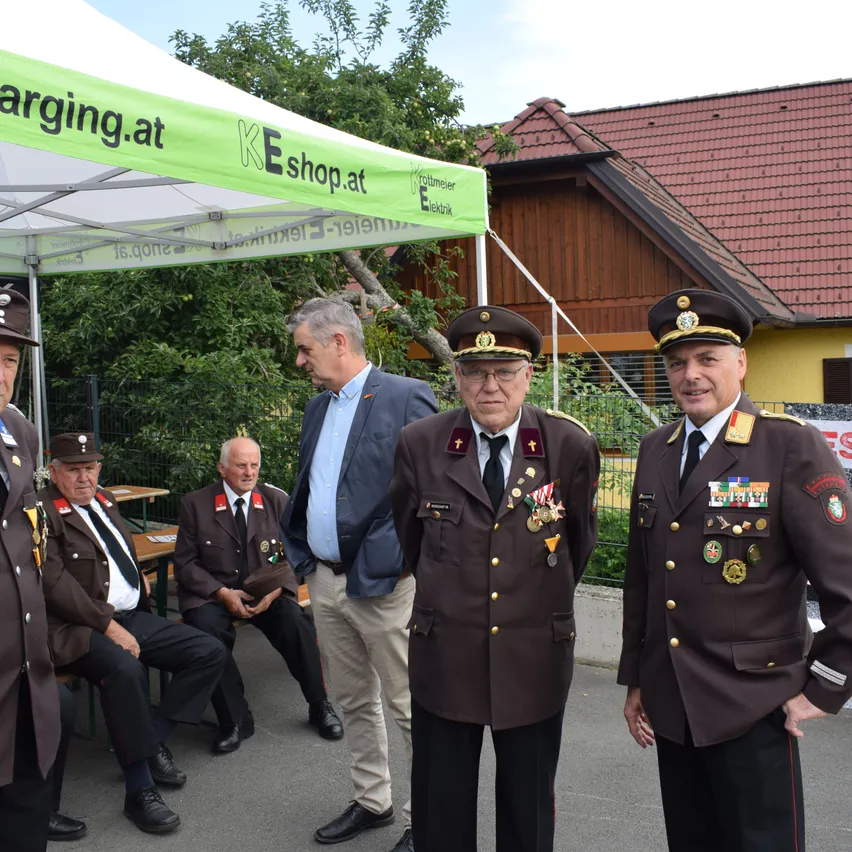 Mehrere Männer in Uniform und ziviler Kleidung stehen vor einem Zelt. Ein Mann in einem Anzug schaut einen sitzenden Mann in Uniform an. Zwei uniformierte Männer stehen neben ihnen.