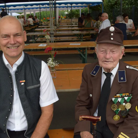Zwei Männer sitzen nebeneinander, einer in einer Weste und der andere in einem Militäruniform mit Medaillen. Dahinter ist eine Gartenparty in vollem Gange, mit mehreren Leuten, die an Tischen sitzen.