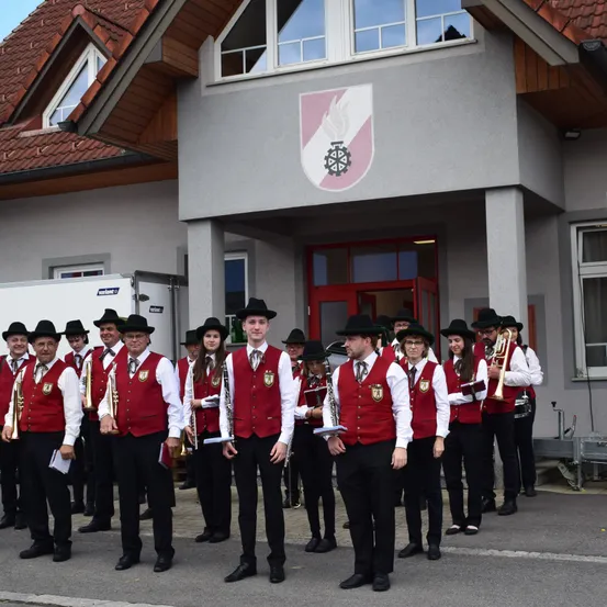 Ein Marschkapelle steht vor einem Gebäude mit Logo. Die Mitglieder tragen rote Westen und schwarze Hüte und halten Instrumente wie Trompeten und Posaunen. Das Gebäude hat Glasfenster und eine rote Tür.