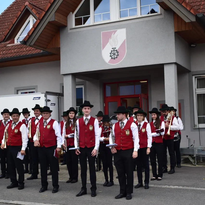 Ein Marschkapelle steht vor einem Gebäude mit Logo. Die Mitglieder tragen rote Westen und schwarze Hüte und halten Instrumente wie Trompeten und Posaunen. Das Gebäude hat Glasfenster und eine rote Tür.