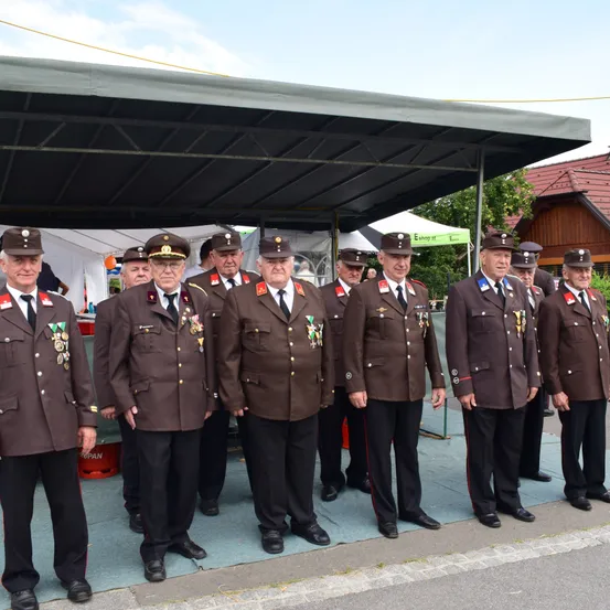 Eine Gruppe von Männern in braunen Uniformen steht unter einem Zelt, trägt Hüte und Medaillen. Dahinter stehen Zelte, Bäume und ein Haus mit rotem Dach. Die Männer stehen in einer Reihe.